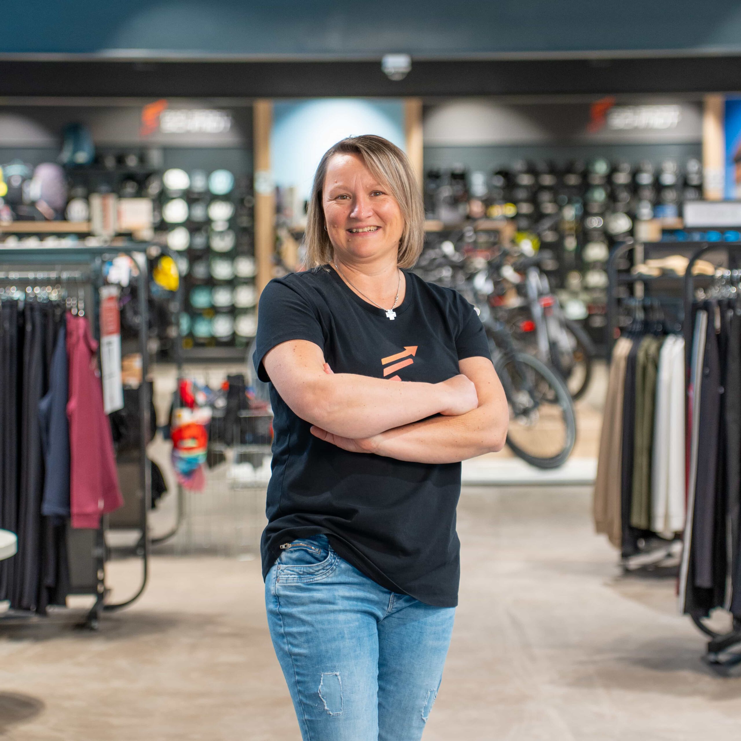 Eine Frau mit kurzen blonden Haaren, die ein schwarzes T-Shirt und blaue Jeans trägt, steht lächelnd und mit verschränkten Armen in einem Geschäft, das Kleidung und Fahrräder ausstellt.