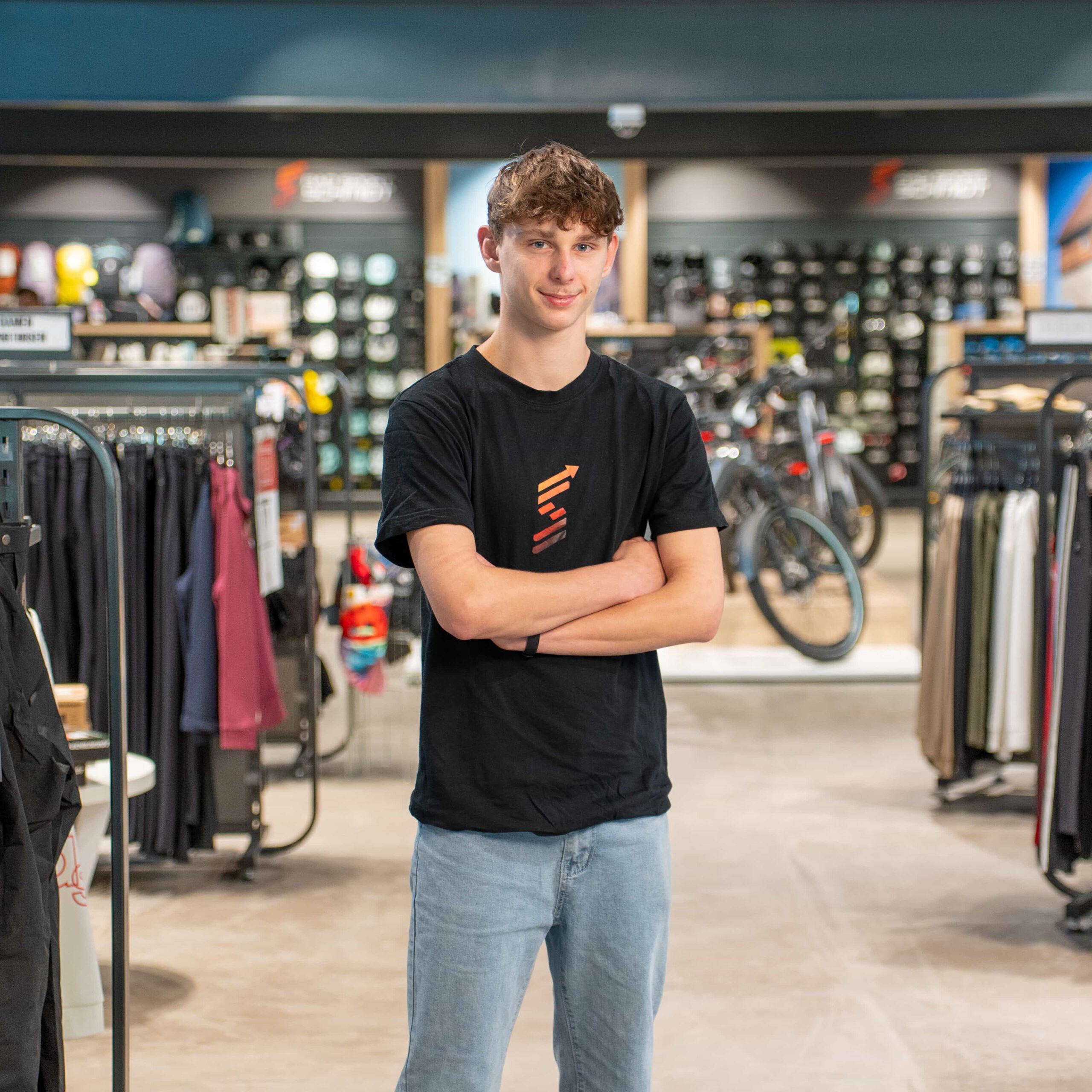 Eine junge Person mit kurzen braunen Haaren, die ein schwarzes T-Shirt und hellblaue Jeans trägt, steht mit verschränkten Armen in einer Ladenpassage, in der im Hintergrund Kleiderständer und Fahrräder zu sehen sind.